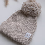 Close-up of a beige knitted baby hat with a fluffy pom pom, textured knit, ribbed folded brim and Snuggle Threads label.