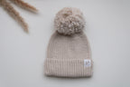 Beige knitted newborn baby hat with a fluffy pom pom, ribbed folded brim and Snuggle Threads woven label.