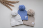 Three knitted baby pom pom hats in ivory, blue and beige, featuring fluffy pom poms, ribbed folded brims and Snuggle Threads woven labels.