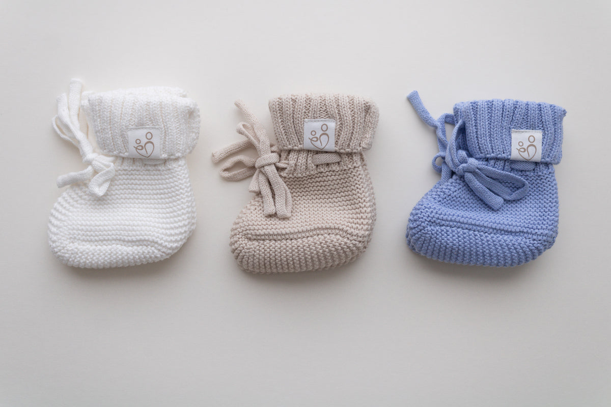 Knitted newborn baby boy booties in ivory, beige and blue, featuring textured knit, ribbed cuffs, tie closures and Snuggle Threads labels.