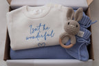 Personalised baby gift box with ivory embroidered romper, blue knitted blanket and booties, and crochet bunny rattle
