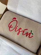Personalised beige knitted baby blanket with red embroidered name and Christmas holly detail, presented in a gift box with white tissue paper