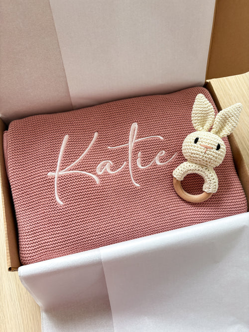 Dusty pink knitted baby blanket personalised with the name Katie in ivory embroidery, presented in a gift box with a crochet bunny rattle.