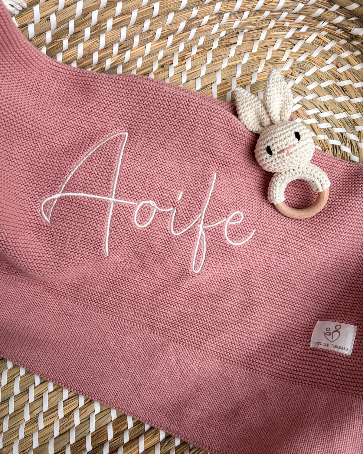 Dusty pink knitted baby blanket personalised with an embroidered name, displayed with a crochet bunny rattle