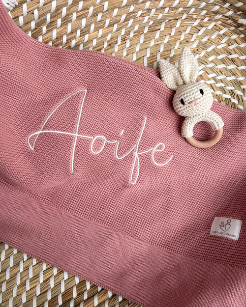 Dusty pink knitted baby blanket personalised with an embroidered name, displayed with a crochet bunny rattle