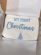 White knitted baby blanket with blue My First Christmas embroidery and Christmas tree design, presented in an open gift box with tissue paper.
