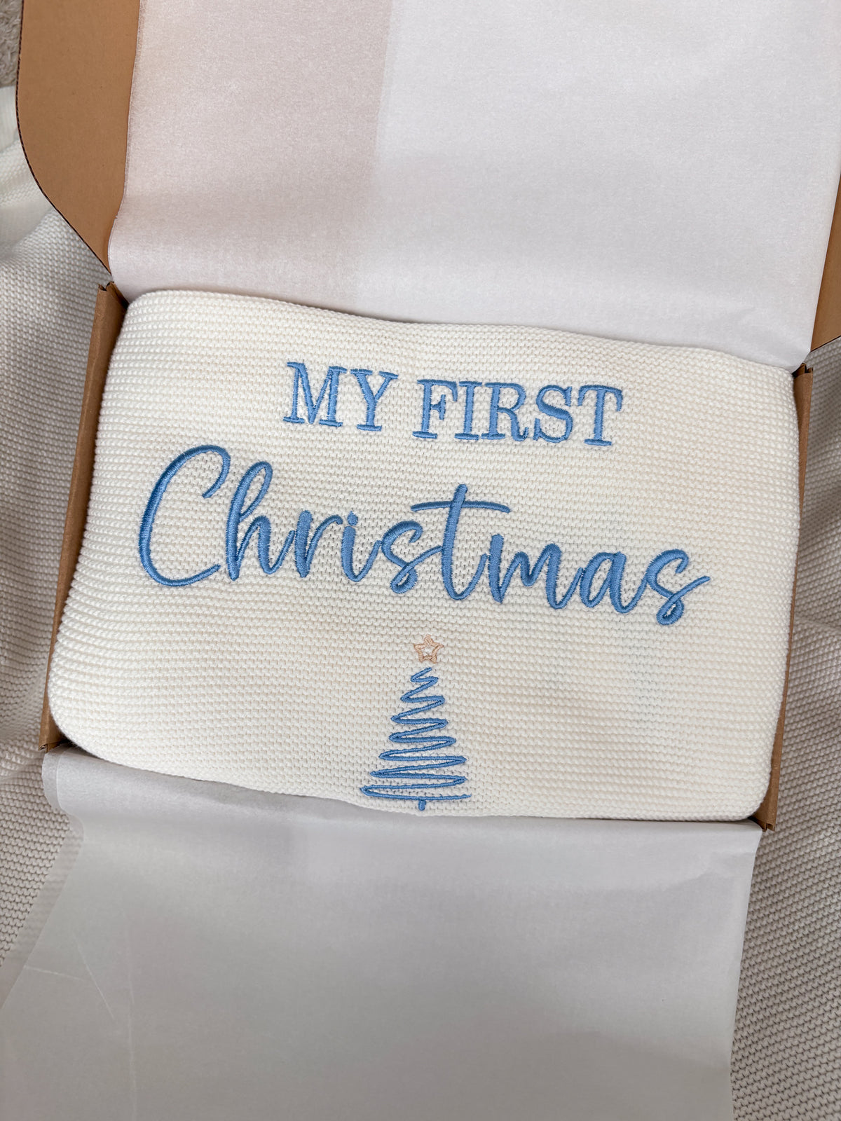 White knitted baby blanket with blue My First Christmas embroidery and Christmas tree design, presented in an open gift box with tissue paper.