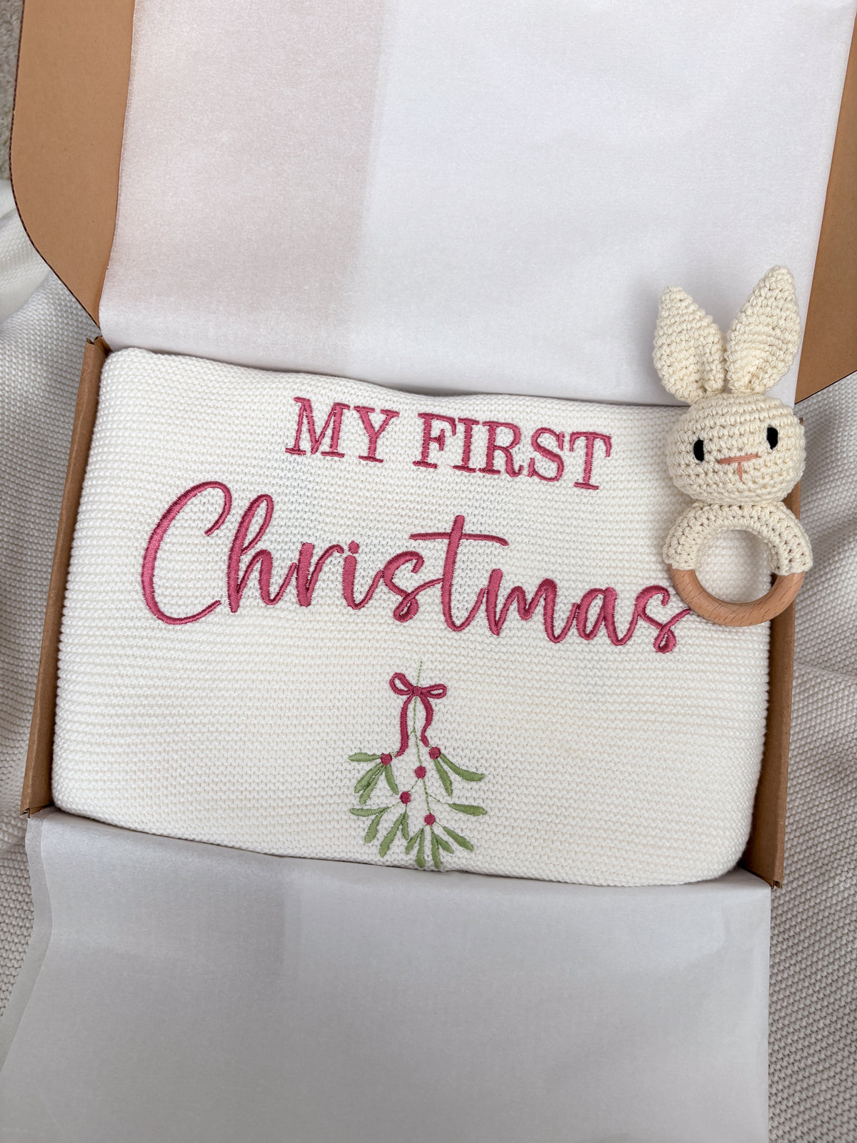 White knitted baby blanket personalised with pink My First Christmas embroidery and mistletoe, presented in a gift box with a crochet bunny rattle.